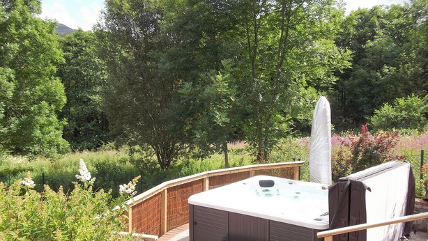 A hot tub with a wooden deck and steps, surrounded by lush greenery and trees.