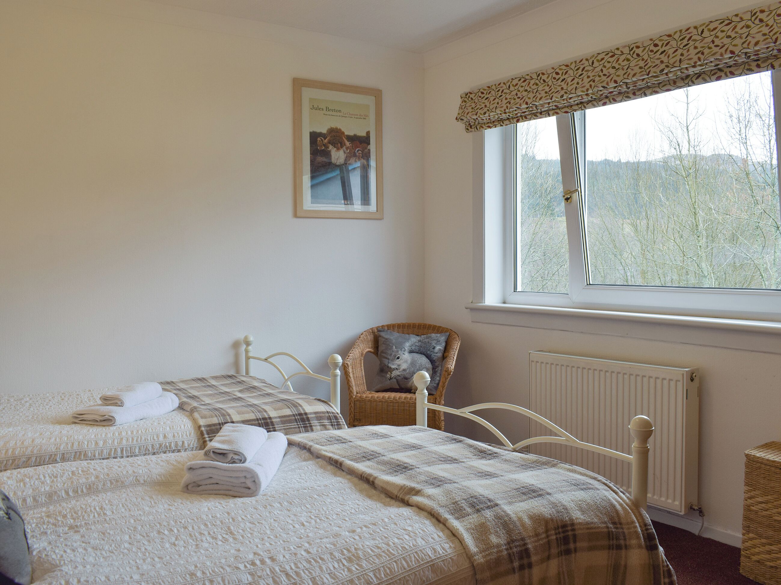 Ein Schlafzimmer mit zwei Betten, einem Korbstuhl, einem Heizkörper und einem Fenster mit Blick auf Bäume.