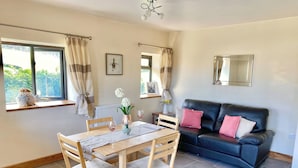 A dining area with a wooden table and chairs, a black leather sofa with pink cushions, and a window with curtains. - Oak View Cottage (Heyope, near Knighton)