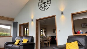 A spacious living room with two black leather sofas, a large wall clock, and a view of a dining area. - Oak View Cottage (Heyope, near Knighton)