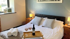 A neatly made bed with pillows, a bedside table with a lamp, a tray with wine glasses and a bottle, and a framed picture on the wall. - Oak View Cottage (Heyope, near Knighton)