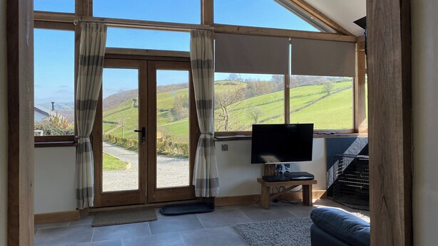 A spacious room with large windows, a television, a small table, and a pet cage. - Oak View Cottage (Heyope, near Knighton)