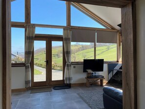 A spacious room with large windows, a television, a small table, and a pet cage. - Oak View Cottage (Heyope, near Knighton)