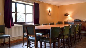 Ein Esszimmer mit einem großen Holztisch, mehreren Stühlen, einem Fenster mit Vorhängen, einer Wandlampe und einem Heizkörper.