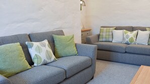 Un salon moderne avec deux canapés gris, des coussins verts ornés de motifs de feuilles, une table basse en bois et une petite table d’appoint avec une lampe.