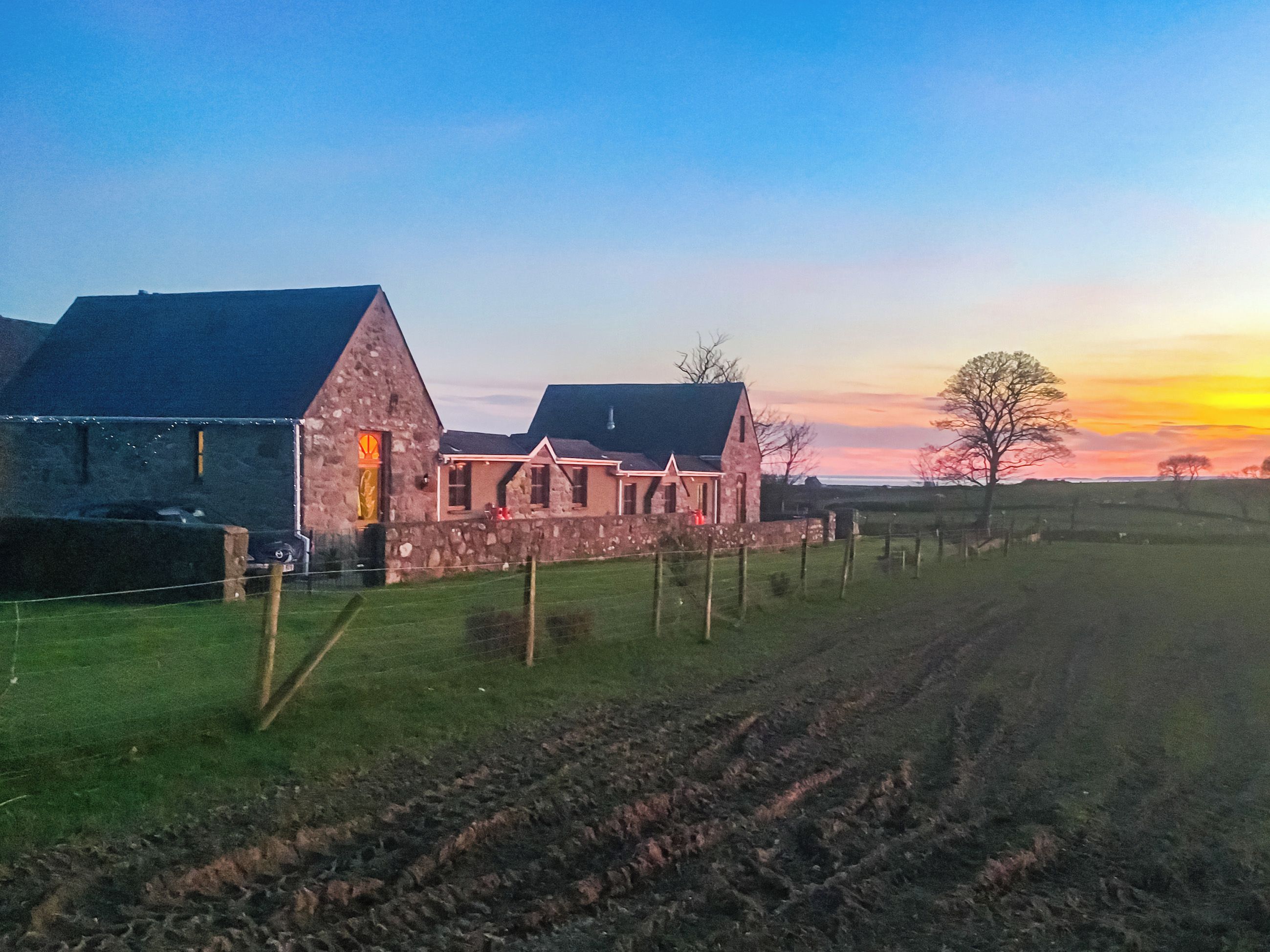 A stone farmhouse with a sloped roof, a fenced yard, and a plowed field.