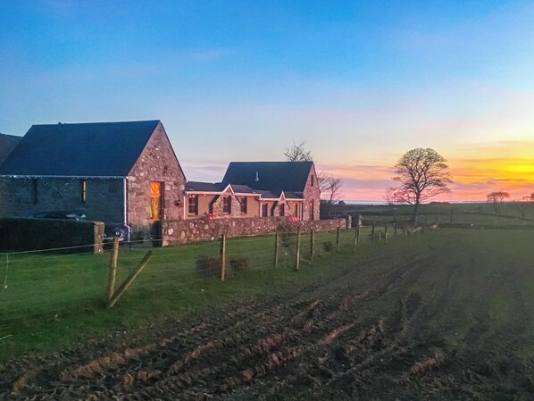 A stone farmhouse with a sloped roof, a fenced yard, and a plowed field.