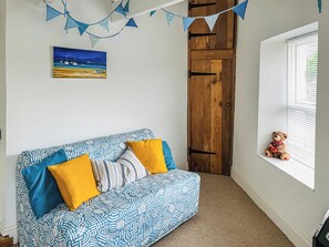 A cozy room with a blue patterned sofa, yellow cushions, a wooden door, a window with blinds, and a painting of a beach scene.