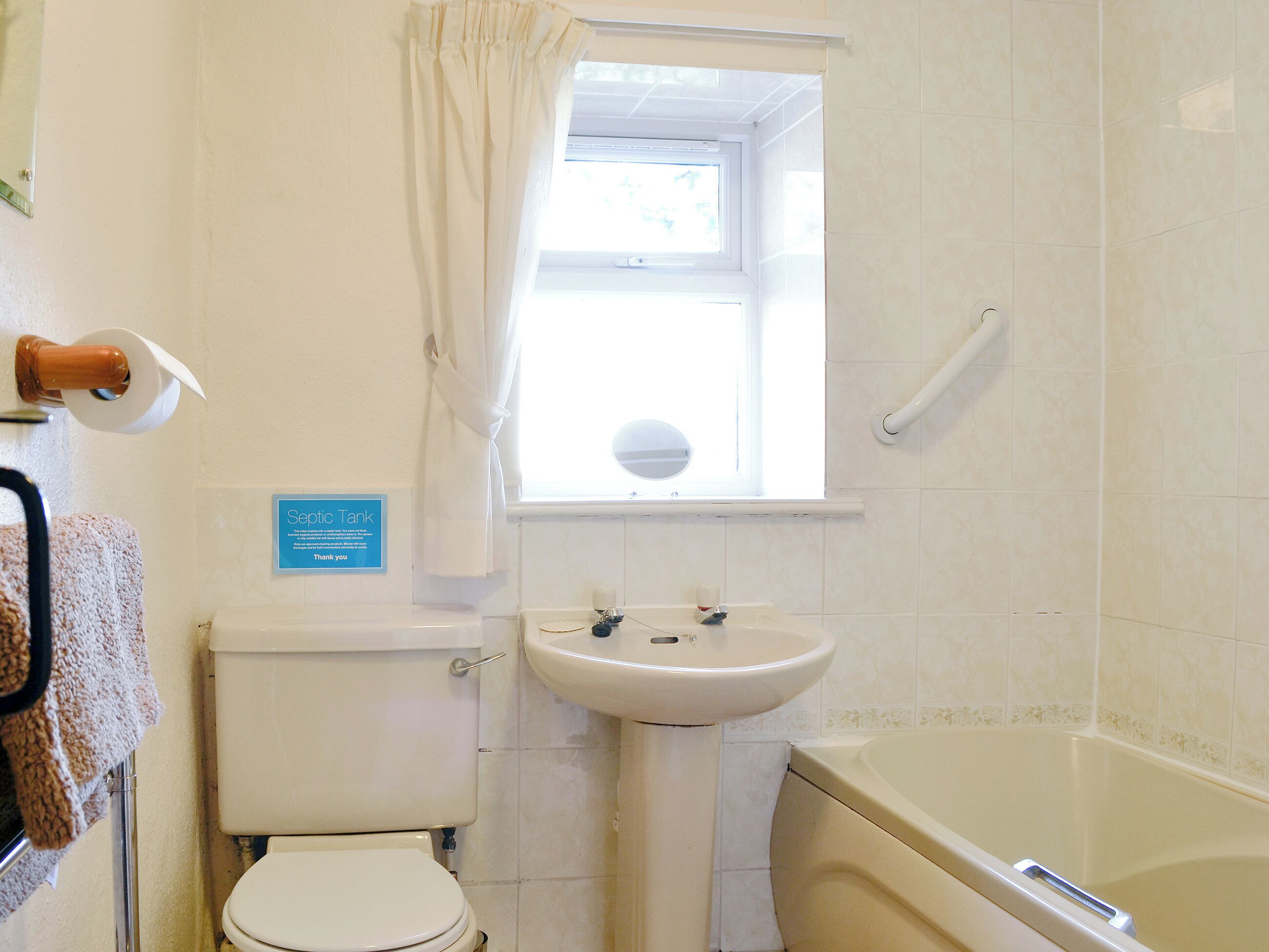 A bathroom with a toilet, sink, and bathtub. There is a window with a curtain, a towel rack, and a sign indicating a septic tank.