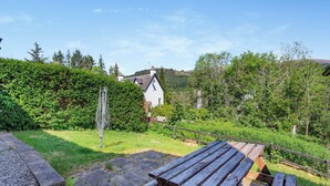 Ein hölzerner Picknicktisch in einem Garten mit einem Steinweg, umgeben von viel Grün und einem weißen Haus im Hintergrund.