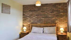 A bedroom with a brick wall, a wooden bed frame, two bedside tables with lamps, and a small wooden dresser.