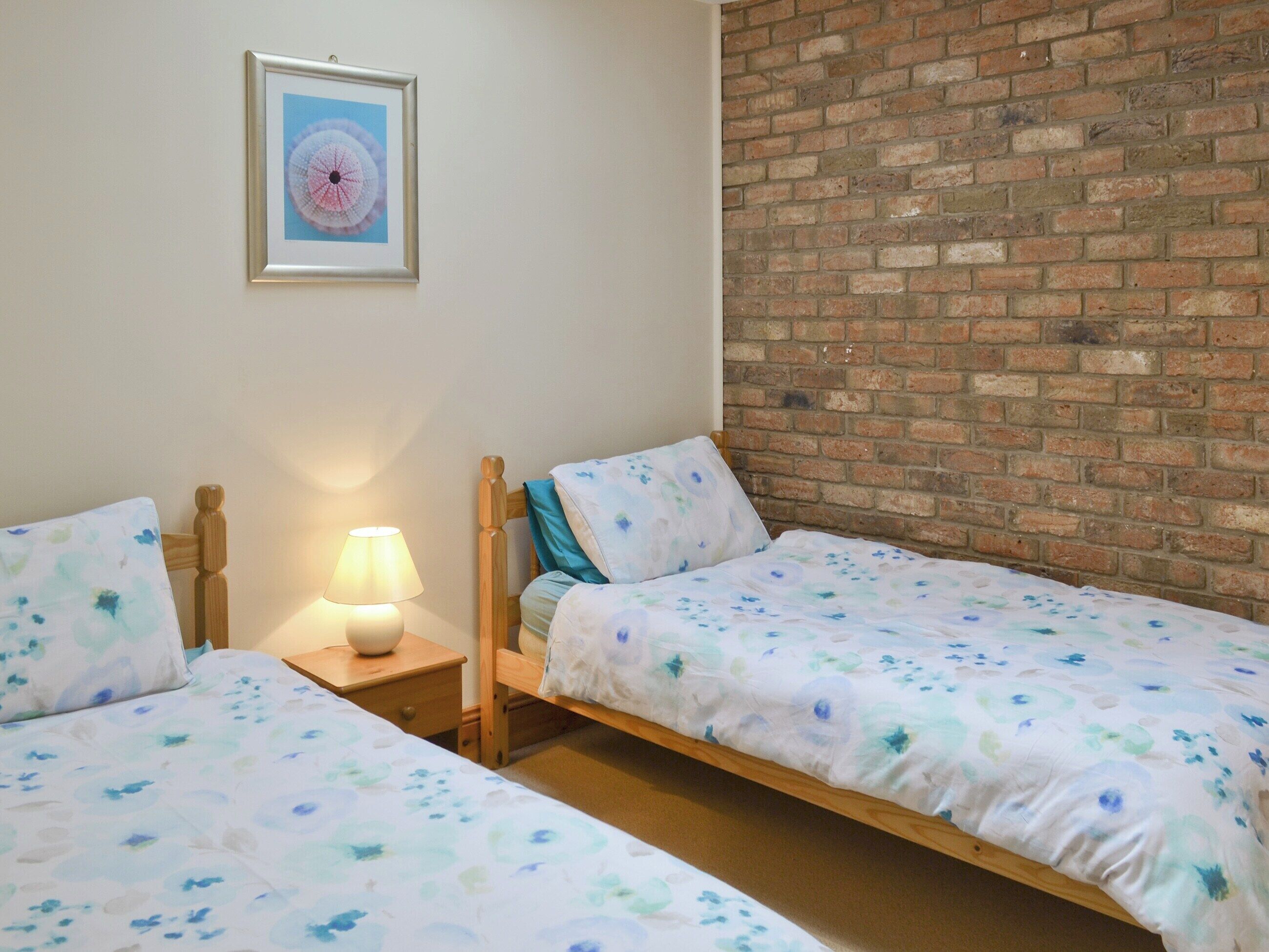 A room with two wooden beds, a nightstand with a lamp, a framed picture on the wall, and a brick wall.