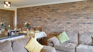 A living room with a brick wall, two sofas, a dining table with chairs, and a kitchen area with a counter and a hanging light fixture.
