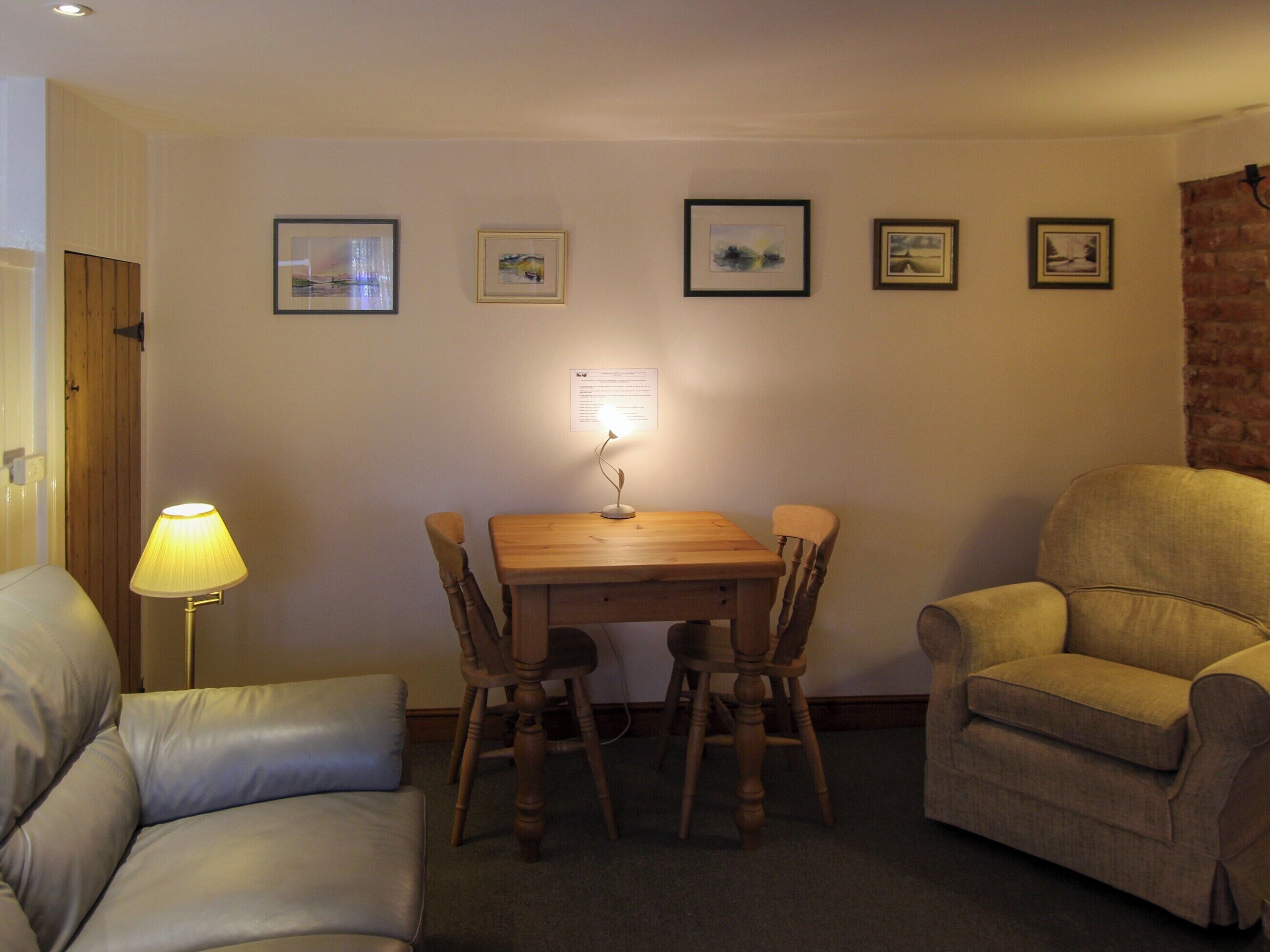 A cozy living room with a wooden dining table, two chairs, a sofa, and a lamp. There are framed pictures on the wall.