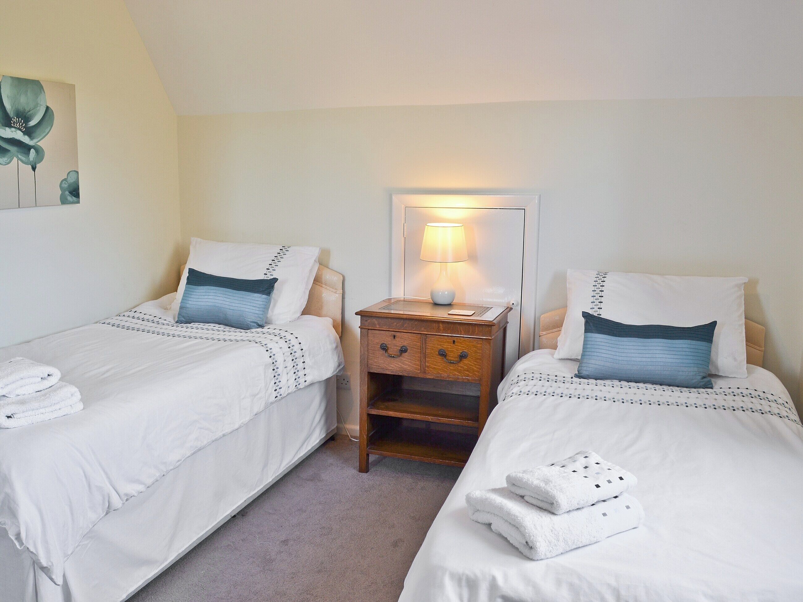 Une chambre d’hôtel avec deux lits simples, chacun équipé d’un oreiller, d’une table de chevet avec une lampe et d’une petite table de nuit en bois.