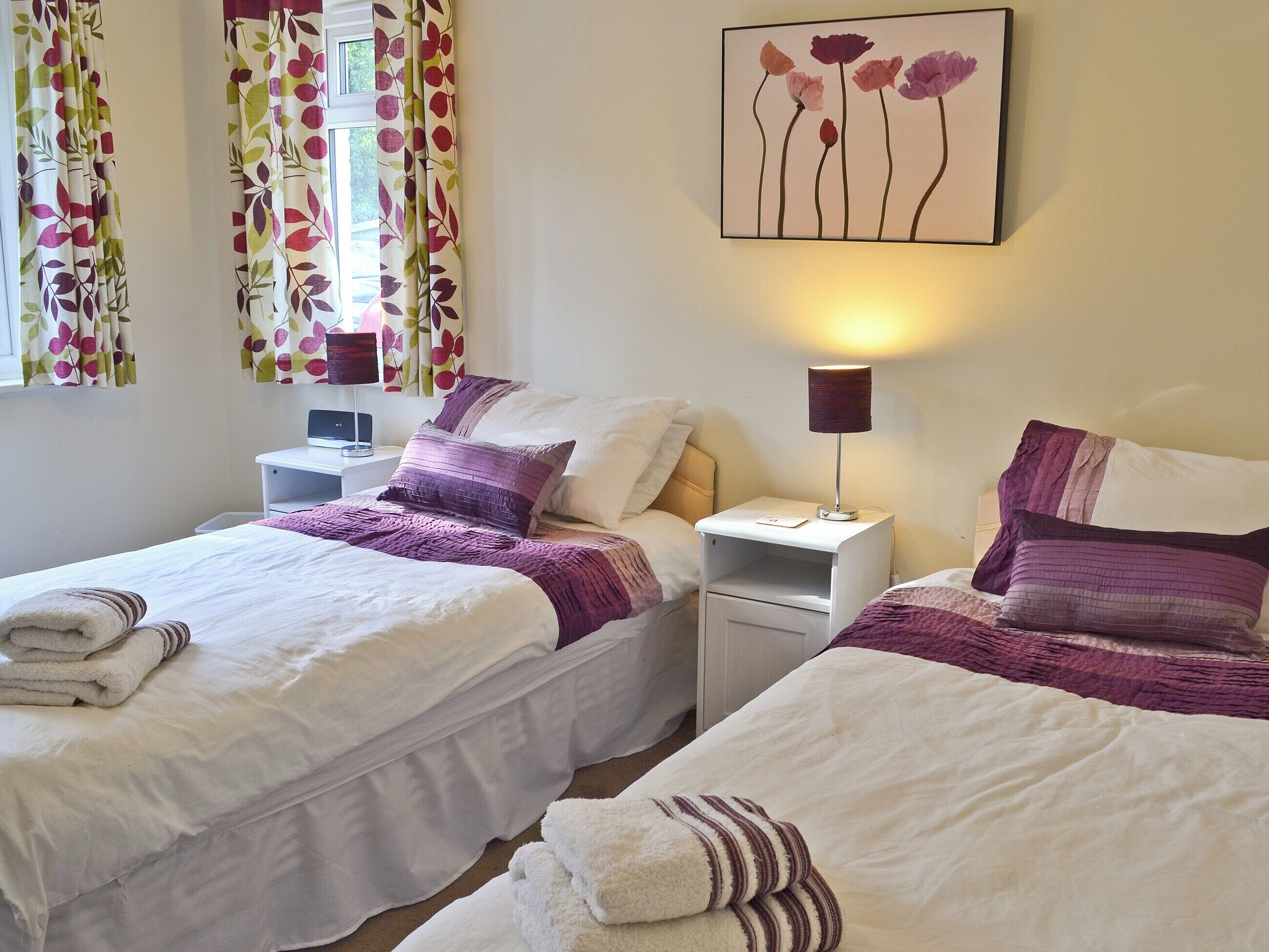 A hotel room with two beds, white bedding, purple accents, a nightstand with a lamp, and a floral artwork on the wall.