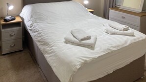 A neatly made bed with white linens and two folded towels on top. There are two bedside tables with lamps, one on each side of the bed.