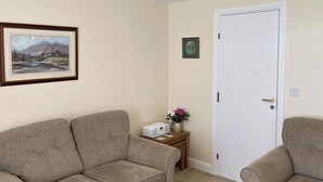 A living room with two beige sofas, a small wooden side table with a vase of flowers, a framed landscape painting, and a white door.