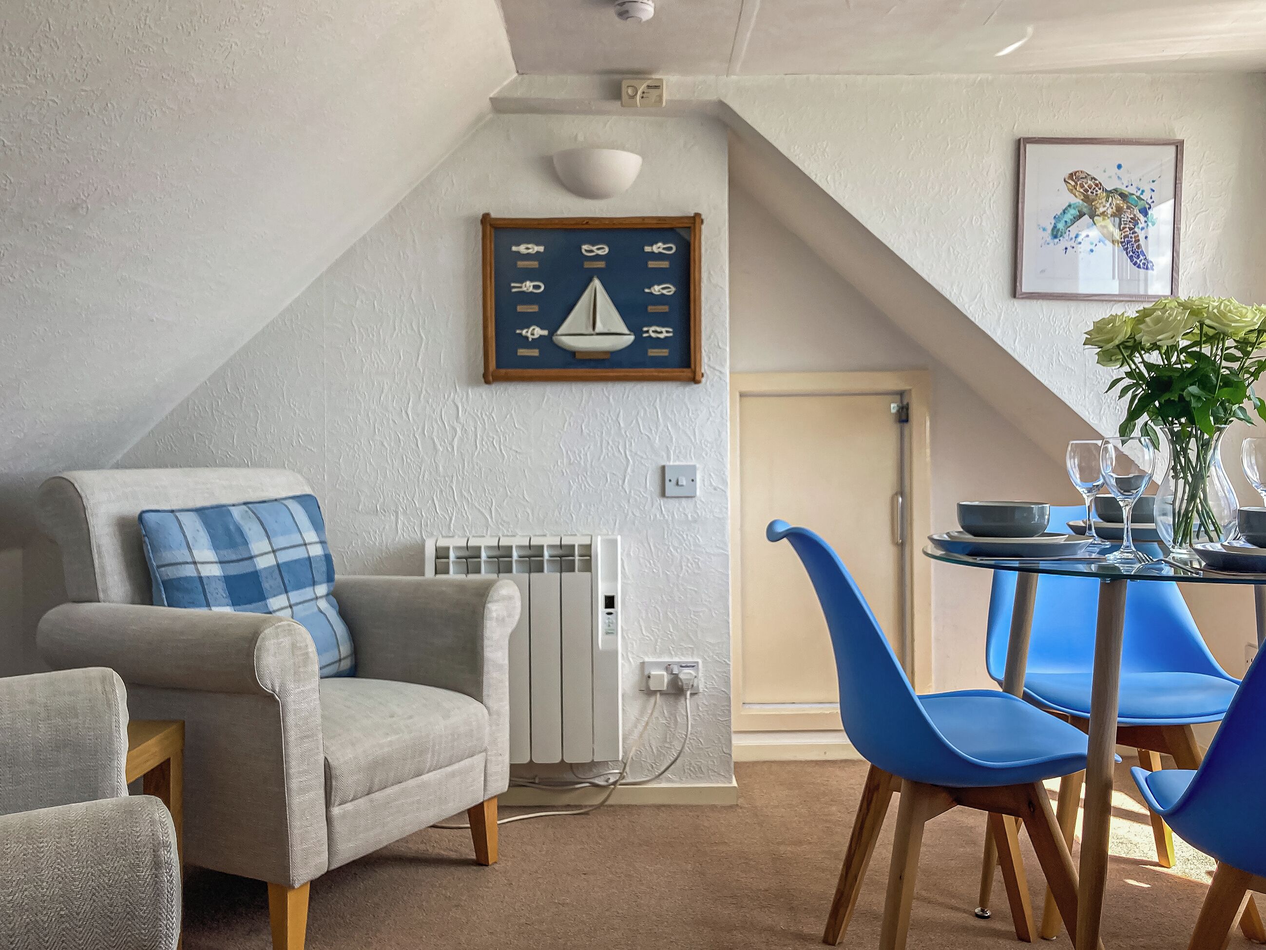 En hyggelig stue med en grå lænestol, en blå sofa, et spisebord med blå stole, en radiator og et billedkunstværk med søfarts tema.