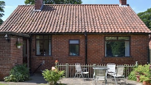 Ein Backsteinhaus mit Ziegeldach, einer kleinen Veranda mit Stühlen und einem Garten.