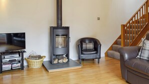 A living room with a black wood-burning stove, a television on a stand, a leather sofa, and a wooden staircase. - 3 bedroom accommodation in Kidderminster (Kidderminster)