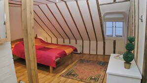 A room with a sloped ceiling, a bed with a red blanket, a small table with a potted plant, and a window with white shutters. - An unusual two-storey apartment with beams and an open fire. (Rye)