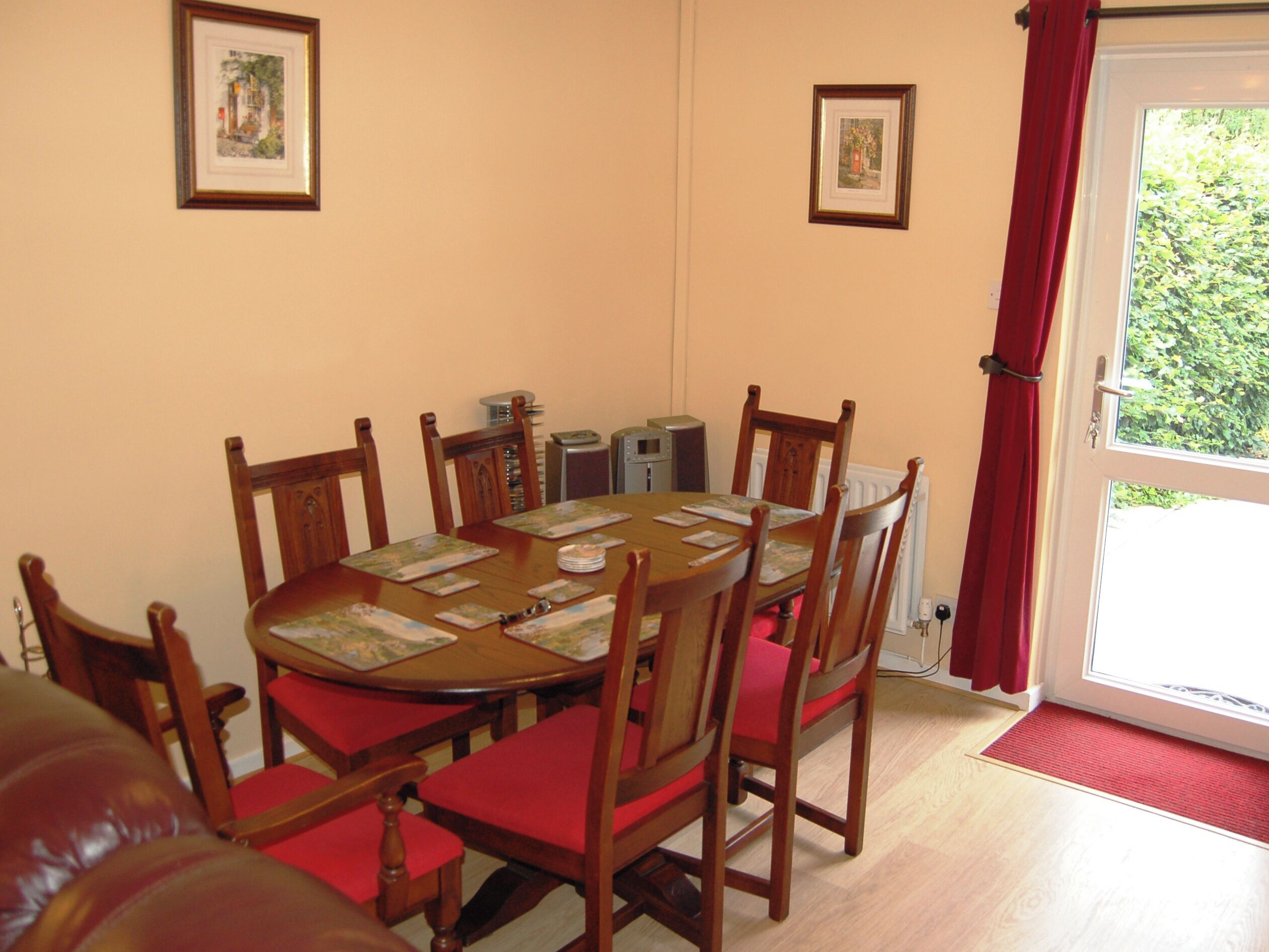 Un coin repas avec une table ronde, des chaises en bois et des coussins rouges. Les murs sont ornés de tableaux encadrés et une fenêtre est agrémentée de rideaux rouges.