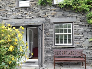 A stone building with a wooden bench outside, a door leading inside, and a window with a grid pattern.