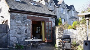 Un bâtiment en pierre avec une porte en bois, un petit patio avec une table et des chaises, et un barbecue.