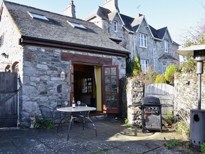 A stone building with a wooden door, a small patio with a table and chairs, and a barbecue grill.