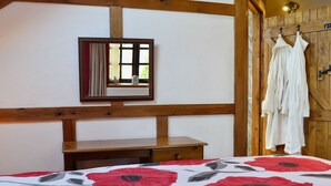 Une chambre avec un plafond en bois, un lit recouvert d’une courtepointe à motifs floraux rouges et gris, une table de chevet en bois, un miroir et des serviettes blanches suspendues à un porte-serviettes en bois.