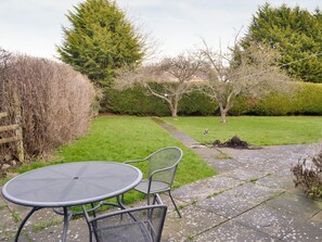 A patio with a round metal table and two chairs, surrounded by a grassy lawn and a hedge.