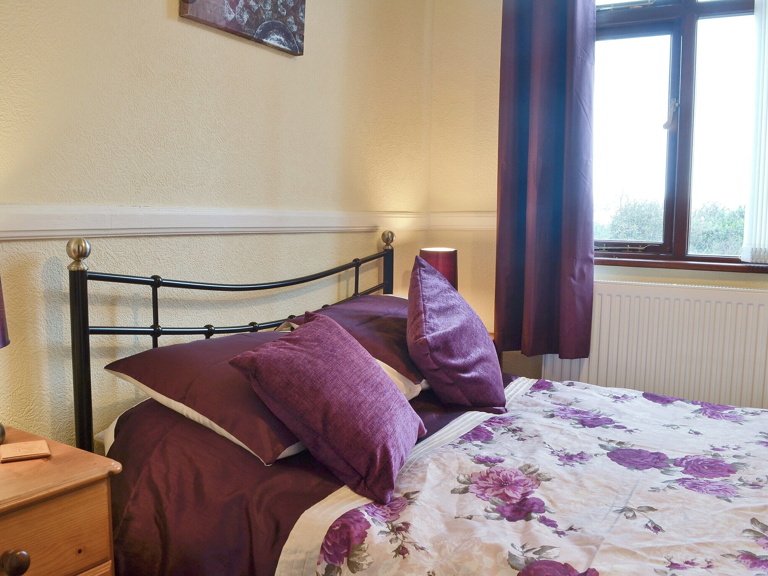 A bedroom with a bed, purple bedding, a wooden nightstand, a wall-mounted lamp, and a window with curtains.