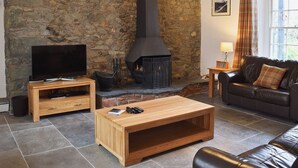 A cozy living room with a stone wall, a fireplace, a television, a leather sofa, and a wooden coffee table. - 4 bedroom accommodation in Keswick (Keswick)