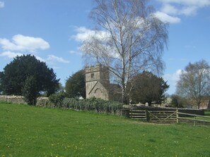 A stone building with a tower, surrounded by a stone wall and a wooden fence, set in a grassy field with trees.