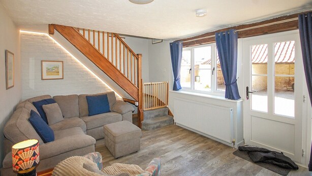 A cozy living room with a wooden staircase, a sofa with cushions, a radiator, and a window with curtains. - 2 bedroom accommodation in Wells-next-the-Sea (Wells-next-the-Sea)