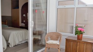 Un dormitorio con una puerta corrediza de vidrio que da a un balcón, una silla de mimbre, un armario de madera y una cama con cubrecama blanco.