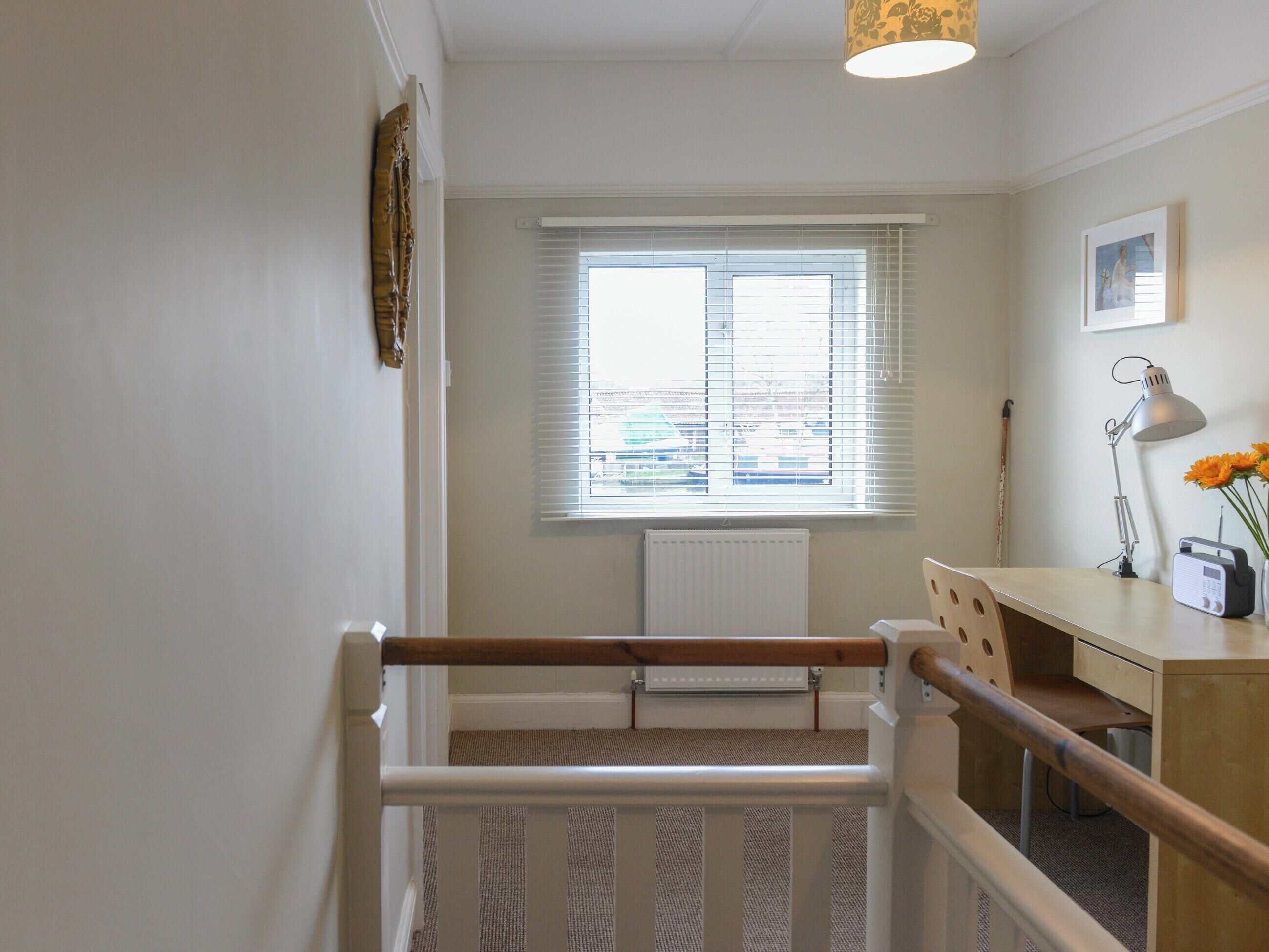 A staircase with a wooden handrail leading to a room with a desk, a radiator, and a window with blinds.