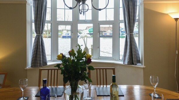 Une table à manger ornée d’un vase de fleurs, de verres à vin et de bouteilles, placée devant une fenêtre à rideaux à carreaux, d’où l’on peut admirer des bateaux sur l’eau.