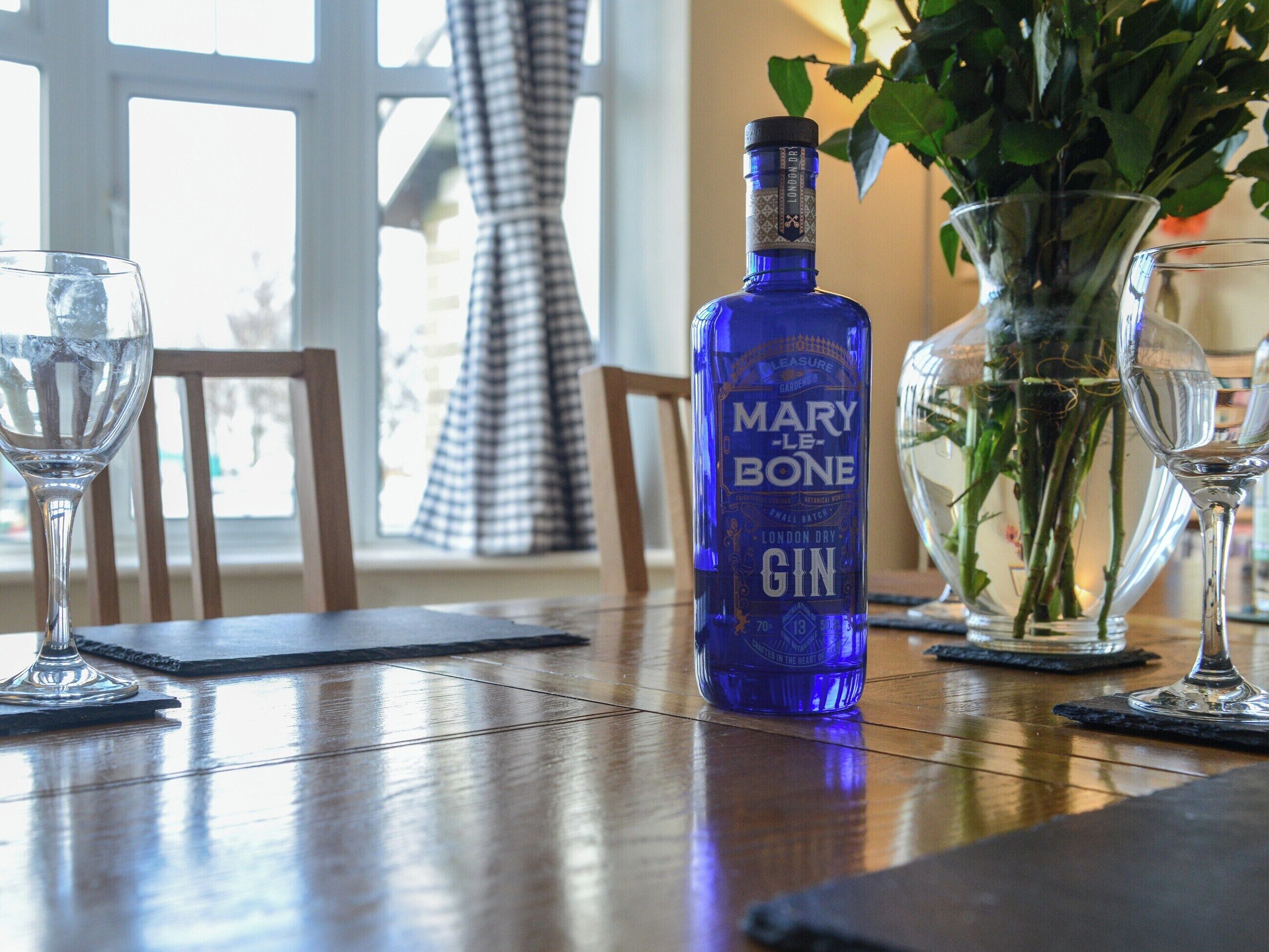 A bottle of Mary Le Bone gin, a vase with green plants, and two empty wine glasses on a wooden table.