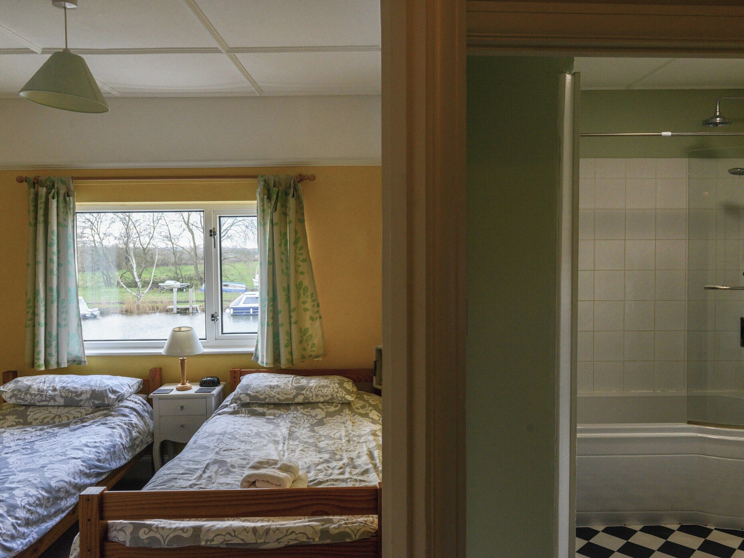 A bedroom with two single beds, a window with curtains, a bedside table with a lamp, and a view of a bathroom with a shower.