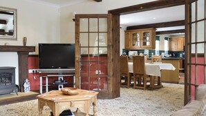 Ein Wohnzimmer mit Fernseher, Holzmöbeln, einem Kamin und einer durch eine offene Tür sichtbaren Küche.