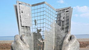 Eine Metallskulptur, die Hände zeigt, die eine strukturierte Darstellung wie eine Karte halten, mit Text und Zahlen, vor einer Küstenlandschaft.