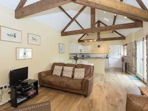 A living room with a brown leather sofa, a TV stand with electronic devices, wooden beams on the ceiling, a kitchen area with white cabinets, and a stone wall. - Westerdale Barn - 27944 (Kildale, near Stokesley)