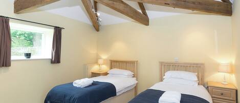 A bedroom with two beds, wooden beams, a window with curtains, bedside lamps, and a view of greenery outside.