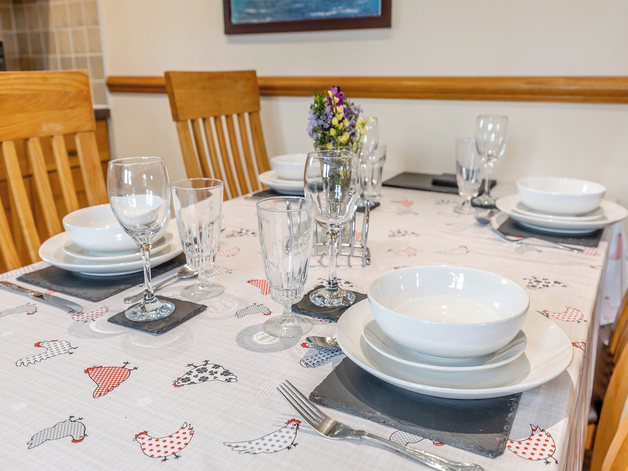 A dining table set with plates, bowls, glasses, and cutlery, with a floral tablecloth and a vase of flowers.