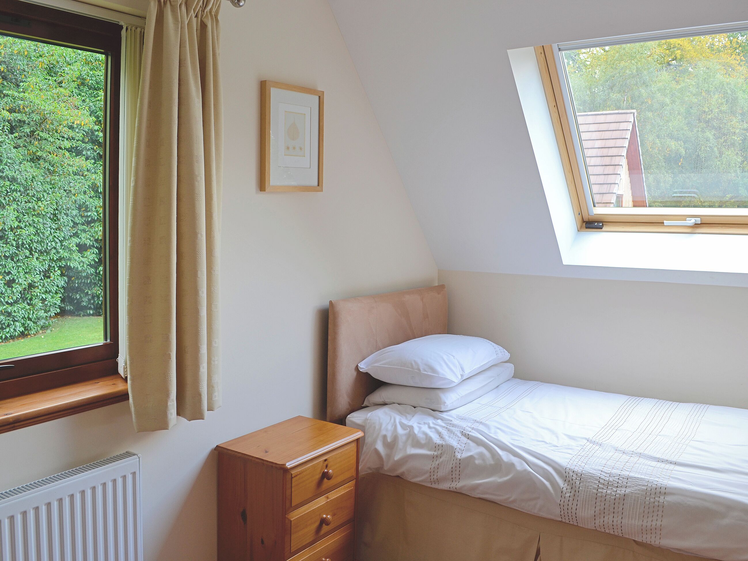 A bedroom with a single bed, a small wooden dresser, a radiator, a window with curtains, and a framed picture on the wall.