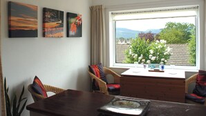 Een eethoek met een houten tafel en stoelen, een raam met uitzicht op groen en bergen, en drie ingelijste kunstwerken aan de muur.