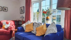 Een woonkamer met een rode fauteuil, een blauwe bank met gestreepte kussens, een bijzettafeltje met een boek en een kopje, en een raam met uitzicht op groen.