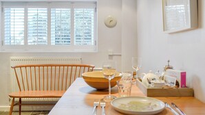 Ein Essbereich mit einem Tisch für zwei Personen, einem Holzstuhl, einer Schüssel, einem Teller mit Essen, Besteck und einer Uhr an der Wand.